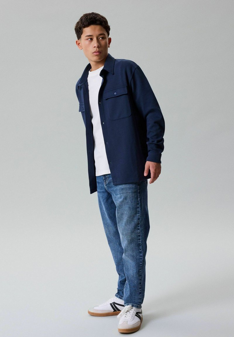 Jeune homme debout regardant de côté, portant une veste-chemise bleu marine, un t-shirt blanc, un jean bleu et des baskets blanches avec des rayures noires.