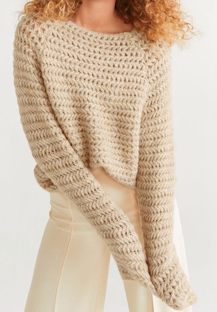 Personne portant un pull épais en maille beige à manches longues et un pantalon ample crème, debout devant un fond clair et uni.