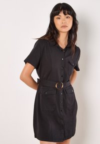 Robe noire à manches courtes, fermée par des boutons à l'avant, avec deux poches poitrine, une taille ceinturée dotée d'une boucle en métal rond et une texture de tissu lisse.