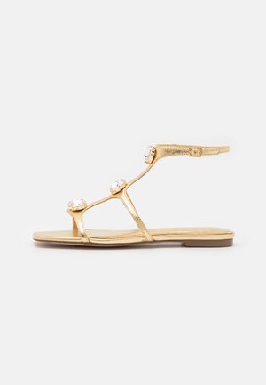 Sandalias planas metálicas doradas con tres gemas transparentes decorativas, correa ajustable en el tobillo y suela lisa. Diseño sencillo con tiras.