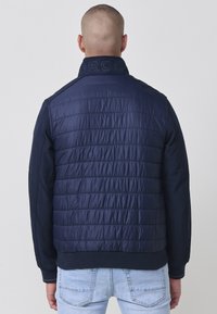 Námornícka puffer bunda s prešívanou textúrou a pevným rukávom. Obsahuje vysoký golier s vytlačeným logom a rebrované manžety a lem.