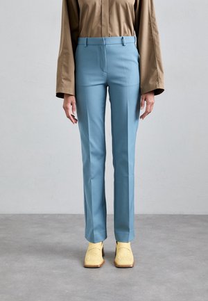 Kobieta w jasnoniebieskich dopasowanych spodniach, beżowej koszuli z długim rękawem i żółtych plecionych botkach na kwadratowym nosku stoi na szarej podłodze.