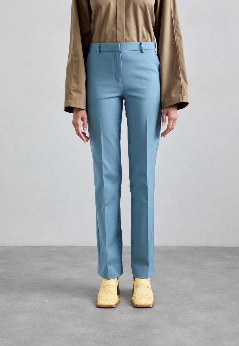 Kobieta w jasnoniebieskich dopasowanych spodniach, beżowej koszuli z długim rękawem i żółtych plecionych botkach na kwadratowym nosku stoi na szarej podłodze.