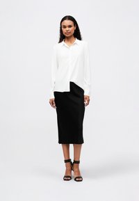 Chemise blanche à manches longues, boutonnée, rentrée dans une jupe midi noire. Le mannequin porte des talons à brides noires sur un fond neutre.