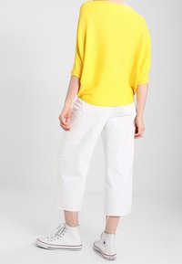 Gelbes geripptes Oversized-Oberteil, weiße cropped Pants und weiße High-Top-Sneaker. Glatter Stoff und lockere Passform mit aufgerollten Ärmeln.