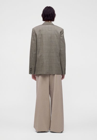 Personne aux cheveux courts et foncés portant un blazer à carreaux et un pantalon large beige, debout de dos sur un fond uni.