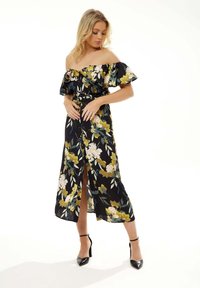 Off-the-shoulder floral dress with ruffled sleeves, black base, multi-colored flowers, front slit, cinched waist, paired with black heels.
