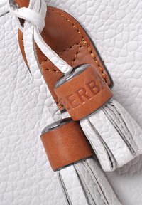 Detail van een witte leren handtas met een bruin leren label en metalen accenten, met een textuuroppervlak en franjes als versiering.