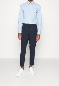 Uomo con una camicia a righe blu chiaro con bottoni, pantaloni tapered navy con coulisse nera e sneakers bianche, in piedi contro uno sfondo bianco.