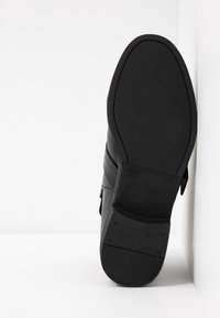 Suela de zapato de cuero negro mirando hacia adelante, con patrón de relieve y marcado de tamaño 37, posicionada en posición vertical contra una pared y un suelo blancos.