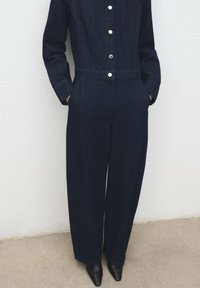 Mono de mezclilla oscura con cintura ajustada, mangas largas, botones frontales y bolsillos laterales. Combinado con zapatos negros puntiagudos.