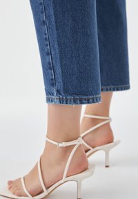Jeans en denim bleu clair de coupe cropped avec une jambe droite. Associés à des sandales à talons hauts blanches à brides avec une fine sangle autour de la cheville.