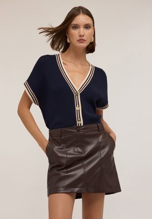 Cardigan corto a maniche corte di colore blu navy in maglia, con scollatura a V, rifiniture in crema e blu scuro, e bottoni dorati, abbinato a una gonna in pelle marrone aderente.