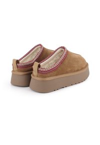 Zapatos slip-on de ante marrón con una gruesa suela de goma beige y un acento de costura roja y blanca alrededor del cuello. Forro suave visible.