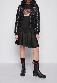 Czarna puchowa kurtka, czarna bluza z kapturem z logo w cętki, plisowana czarna spódnica, czarne botki za kostkę z czerwonym akcentem. Gładkie i fakturowane materiały.