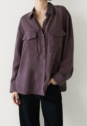 Osoba ubrana w luźną, długą, fioletową koszulę na guziki z kieszeniami na piersiach oraz czarne spodnie, stojąca na jednolitym tle.