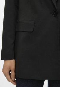 Black tailored blazer with a single button closure, side pocket, and smooth fabric texture; worn over dark denim.