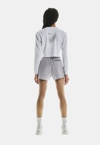 Avklippt långärmad vit skjorta med grafik på baksidan, matchad med ljusgrå shorts med en dragkedjeficka. Modellen bär vita strumpor och skor.