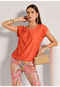 Oranges Kurzarmoberteil mit Falten_detail, kombiniert mit gemusterten Hosen in pink, rot und schwarz, und einer geflochtenen Handtasche.