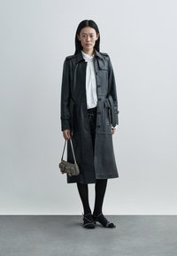 Czarny skórzany płaszcz trencz zapinany na guziki, z długimi rękawami i kieszeniami po bokach, w zestawieniu z białą koszulą, czarnymi spodniami i teksturowaną torebką.