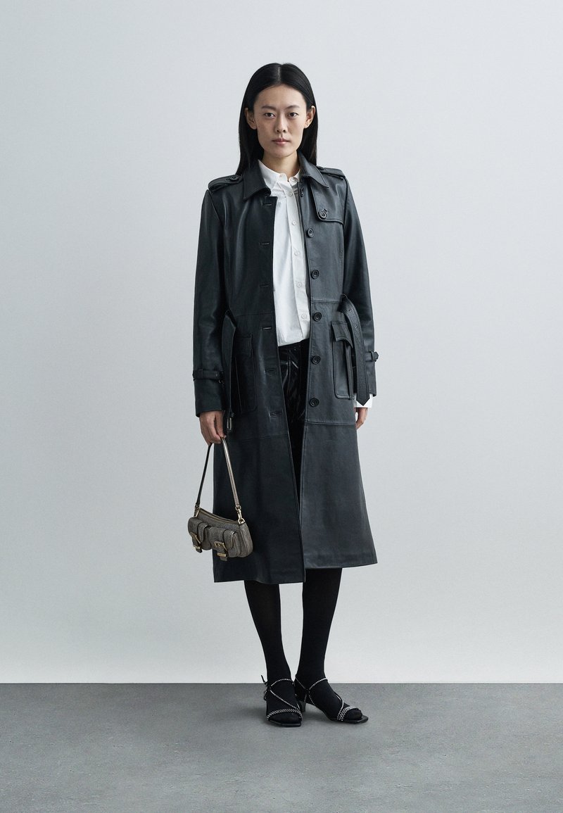 Czarny skórzany płaszcz trencz zapinany na guziki, z długimi rękawami i kieszeniami po bokach, w zestawieniu z białą koszulą, czarnymi spodniami i teksturowaną torebką.