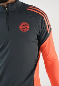 Sudadera deportiva gris y naranja con cuello de media cremallera, que presenta el logo del FC Bayern Munich y tres stripes naranjas en el hombro izquierdo.