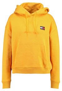 Sudadera amarilla de tela suave, con un bolsillo frontal, capucha con cordón y un pequeño parche de logo con acentos en rojo, blanco y azul.