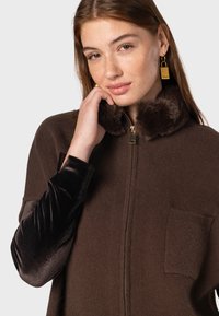 Pull en brown zippé avec un col en fausse fourrure, doté de longues manches en velours noir et d'une poche avant. Texture lisse avec des accents brillants.