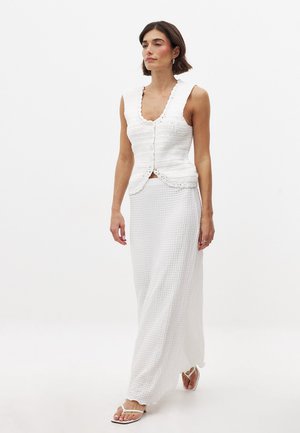 Top court en tricot blanc avec des bords festonnés, présentant un col en V et des boutons, associé à une longue jupe blanche texturée.