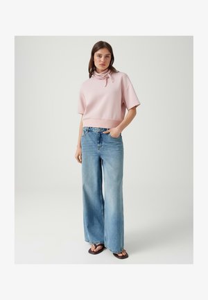 Junge Frau trägt einen hellrosa kurzärmligen Pullover, weite blaue Jeans und schwarze Sandalen und steht vor einem schlichten weißen Hintergrund.