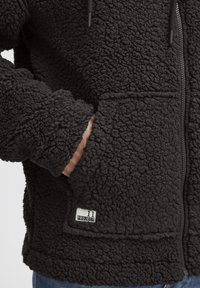 Black fleece jacket with a textured finish, zip closure, and front pouch pocket. Features a small label reading "11 PROJECT" near the hem.