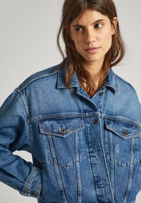 Veste en denim bleu au design classique, dotée de deux poches poitrine, de fermetures à boutons et de détails de couture visibles. Le tissu a une finition texturée.