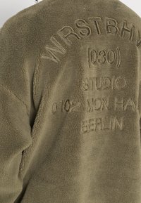 Giacca in pile verde oliva con testo in rilievo: "WRSTBHVM (D30) STUDIO 0102 MON HAI BERLIN." Texture morbida con design casual.