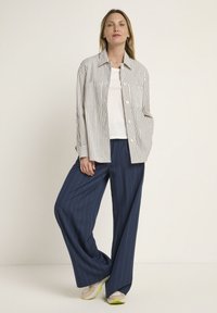 Striped button-up shirt in beige and navy, worn over a white top. Loose navy pinstripe pants. Athletic shoes with green accents.