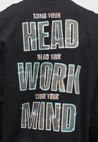 Schwarzer Pullover mit auffälligem, stilisiertem weißen und blauen Grafiktext: "MIND YOUR HEAD, READ YOUR WORK, WORK YOUR MIND."