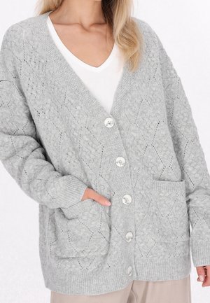 Femme portant un cardigan en maille gris clair avec fermeture par boutons, motif losange texturé, poches avant et poignets côtelés sur un t-shirt blanc col V.
