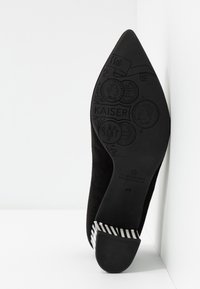 Chaussure à talon haut en daim noir avec un bout pointu, dotée d'une semelle texturée et d'un accent rayé sur le talon. Logo gravé sur la semelle.
