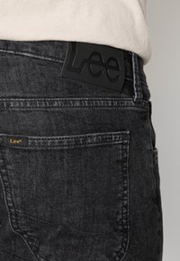 Jeans de denim preto com um acabamento mate. Apresenta um patch de couro com logotipo em relevo e detalhes em laranja perto do bolso traseiro.