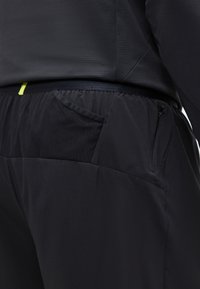 Κοντινό πλάνο μαύρου αθλητικού παντελονιού με ελαστική μέση, δίχτυ στην πλαϊνή τσέπη και τσέπη με φερμουάρ στο ισχίο, φορεμένο με μαύρο κορμάκι.