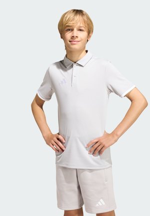 Ragazzo biondo in piedi con le mani sui fianchi indossa una polo Adidas grigio chiaro e pantaloncini abbinati su sfondo neutro.