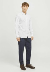 Camisa blanca de manga larga con botones y cuello, combinada con pantalones azul oscuro y zapatos de cordones marrones, de pie contra un fondo neutro.