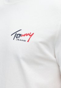 Λευκό ύφασμα που δείχνει το λογότυπο Tommy Jeans με το "Tommy" σε μαύρη και κόκκινη γραφή πάνω από το "JEANS" με μαύρα κεφαλαία γράμματα.