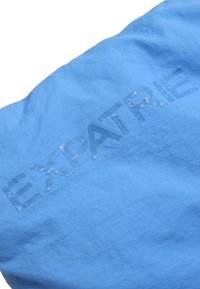 Svetlomodrá látka so štruktúrovaným strieborným potlačou slova "EXPATRIÉ" cez stred, s jemnou a hladkou textúrou.