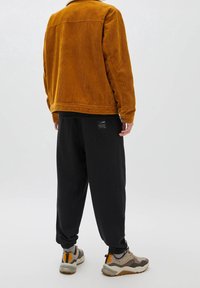 Giacca in corduroy di colore arancione bruciato, vestibilità rilassata, con colletto. Joggers neri con polsini elasticizzati, etichetta accentuata sul retro, abbinati a sneakers chunky.