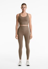Bruine crop top met brede bandjes en bijpassende hoge taille legging; beide zijn gemaakt van soepel, rekbaar materiaal. Witte sneakers maken de look af.