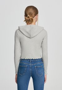 Felpa grigia corta con cappuccio e orlo ondulato, abbinata a jeans blu aderenti con tasche posteriori. Tessuto morbido e a costine.