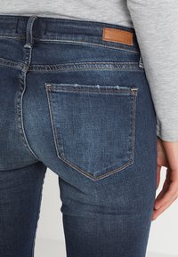 Mörkblå denimjeans med en figursydd design, med en bakficka och en brun läderetikett vid midjan. Slät textur.