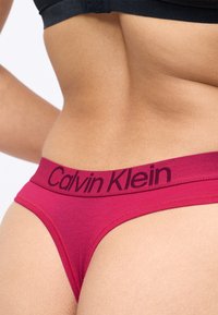 Czerwona bawełniana stringi z czarnym elastycznym paskiem z logo "Calvin Klein". Zbliżenie pokazujące strukturę materiału i dopasowanie.