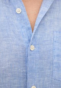 Camicia azzurra chiara in lino, con colletto, tre bottoni, tessuto testurizzato e una tasca sul petto sul lato sinistro.
