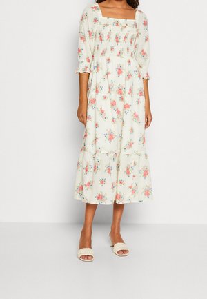 Robe midi florale en crème avec des motifs roses et bleus, présentant un décolleté carré, un corps smocké et un ourlet à volants. Portée avec des sandales neutres.